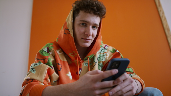 A fashion studio shootingt of a young man in hoodie with smartphone posing over orange background.