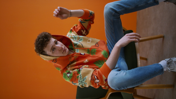 A vertical footage of fashion studio shooting of a young man in hoodie with big leaf sitting in armchair.