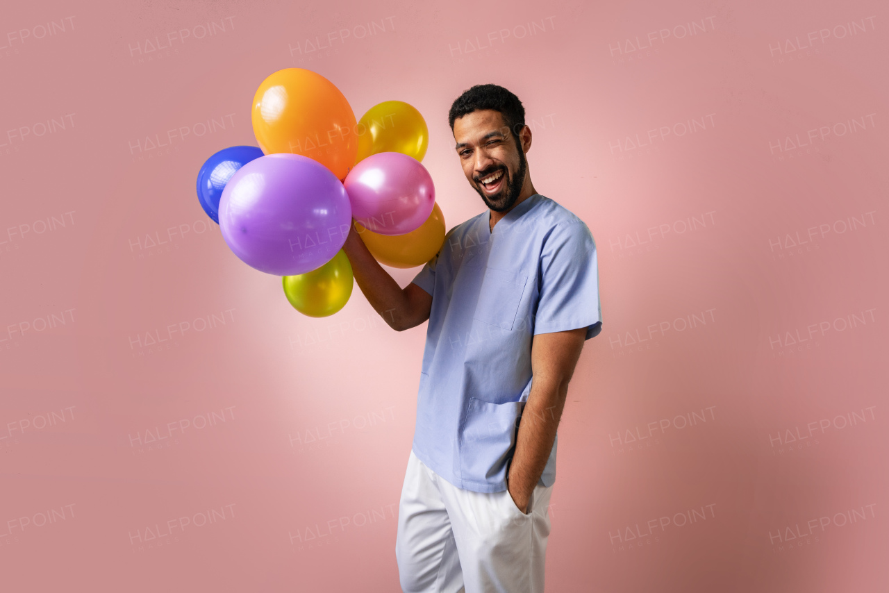 A happy young pediatrician holding balloons over pink background