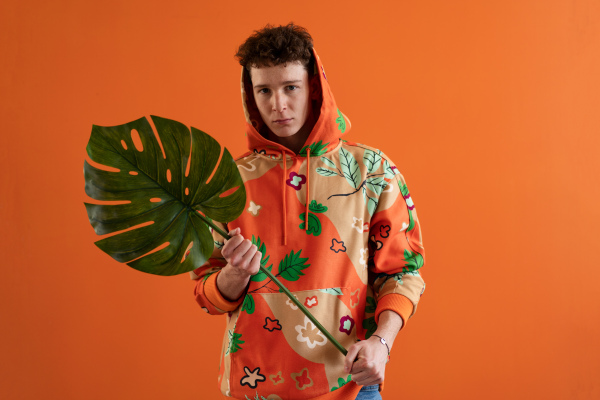 A fashion studio portrait of a young man in hoodie with big leaf posing over orange background.