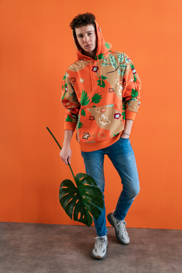 A fashion studio portrait of a young man in hoodie with big leaf posing over orange background.