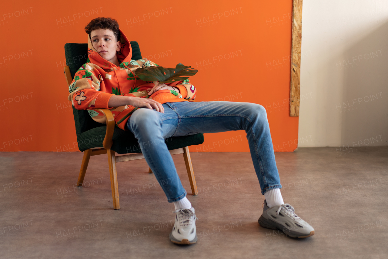 A fashion studio portrait of a young man in hoodie with big leaf sitting in armchair.