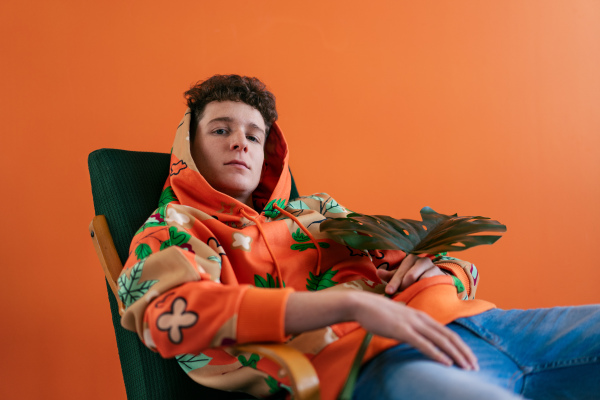 A fashion studio portrait of a young man in hoodie with big leaf sitting in armchair.