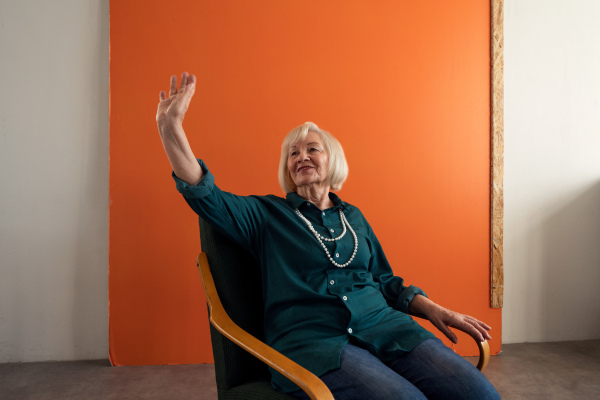 A smiling elegant elderly woman sitting in armchair and waving to someone.