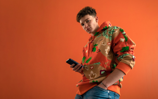 A fashion studio portrait of a young man in hoodie with smartphone posing over orange background.