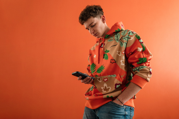 A fashion studio portrait of a young man in hoodie with smartphone posing over orange background.