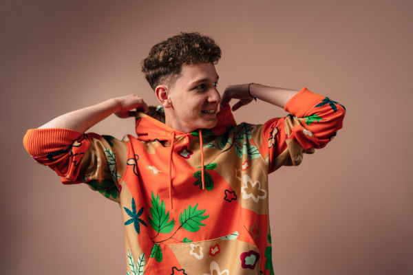 A fashion studio portrait of a young man in cool orange hoodie posing over beige background.