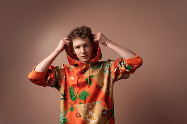 A fashion studio portrait of a young man in cool orange hoodie posing over beige background.