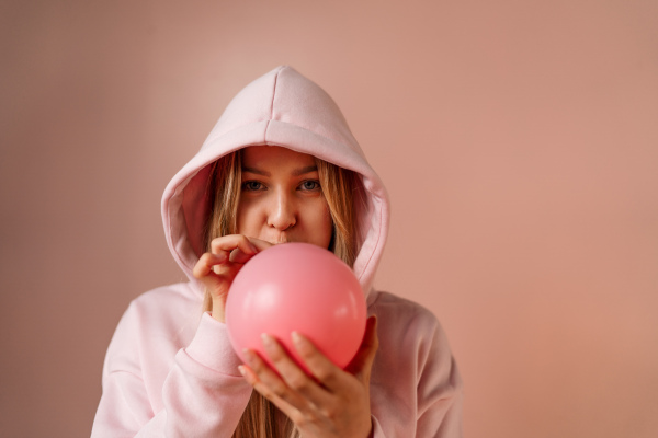 A fashion studio portrait of a happy young blonde woman in hoodie blowing balloon over pink background.