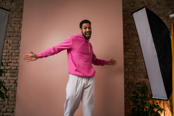 A fashion studio portrait of a young African American man in pink hoodie posing over beige background.