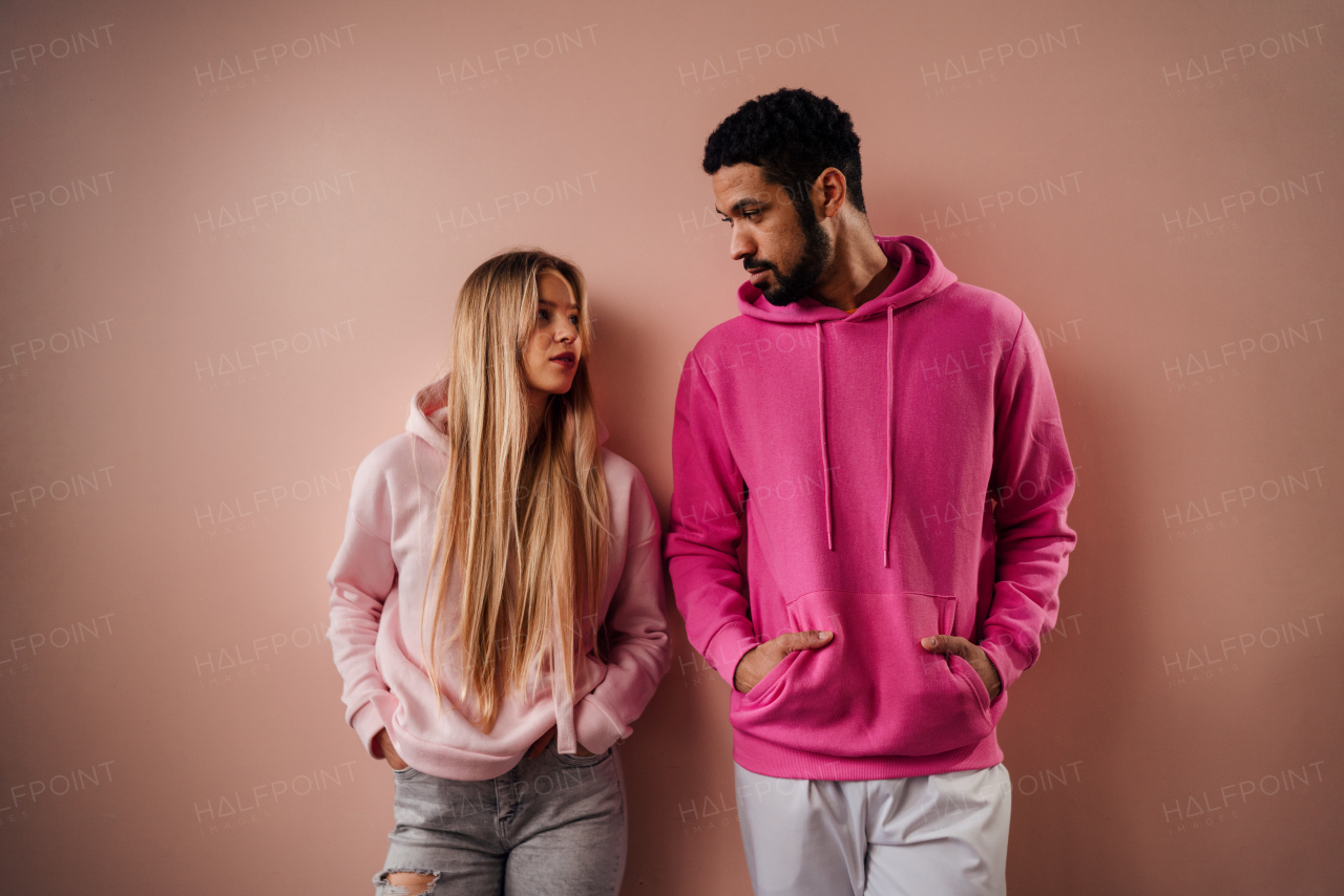 Fashion studio portrait of young couple in hoodie looking at each other and posing over pink background.