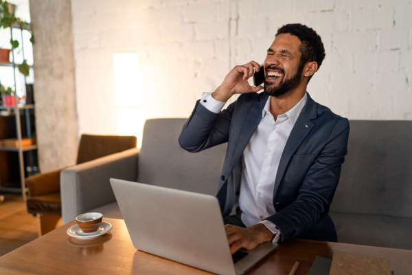 A cheerful young businessman sitting on sofa and talking on cellphone in office.