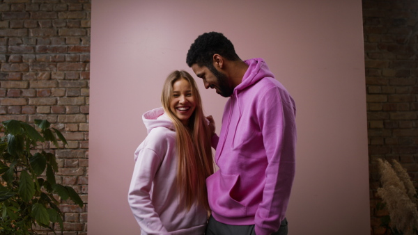 A fashion studio shooting of a happy young couple in hoodie posing over pink background.