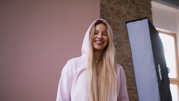 A fashion studio shooting of a happy young blonde woman in hoodie posing over pink background.