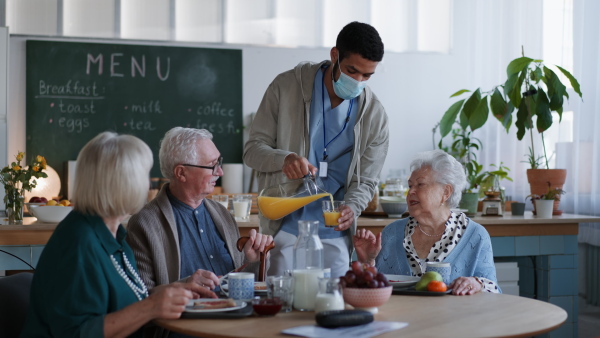 A young caregiver serving breakfast to group of seniors in nursing home care center.