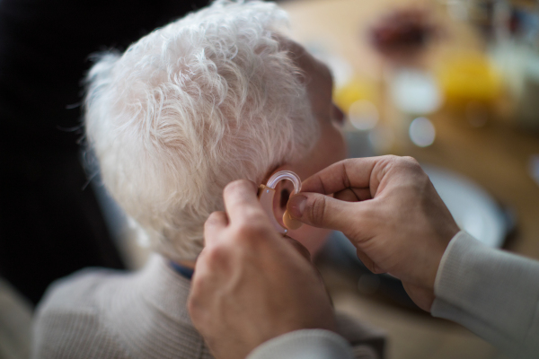 A close-up of caregiver man's hand inserting hearing aid in senior's man ear.