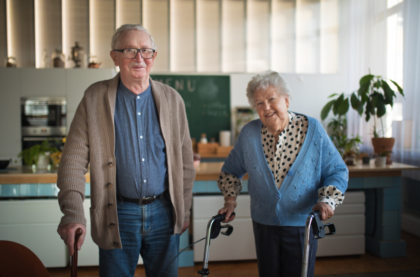 A senior man and woman walking with walker indoors in retirement nursing hme.