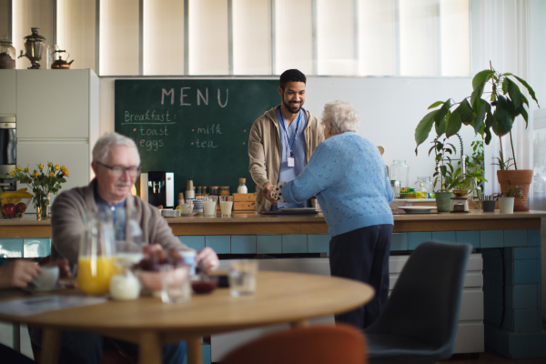 A young caregiver serving breakfast to elderly woman in nursing home care center.