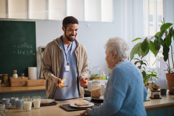 A young caregiver serving breakfast to elderly woman in nursing home care center.