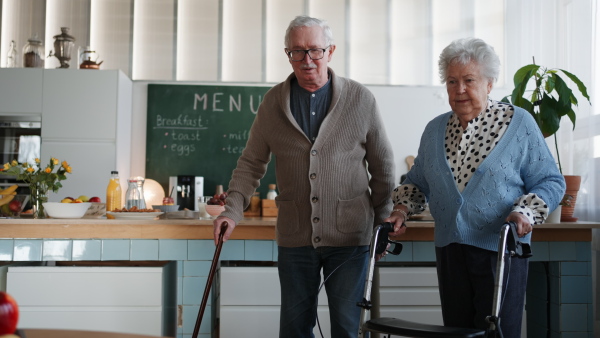 A senior man and woman walking with walker indoors in retirement nursing hme.