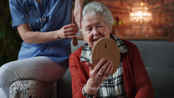 Nurse combing hair of senior woman observing herself in a mirror in retirement home