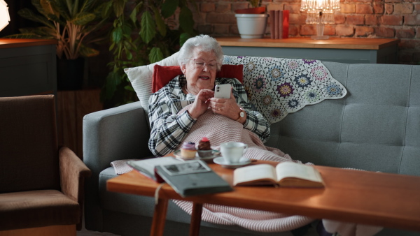 A happy senior woman sitting on sofa at home and using smartphone.