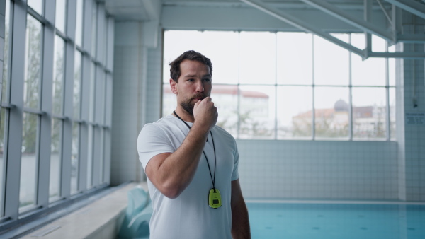 A personal coach whistling and giving instructions indoors in swimming pool.