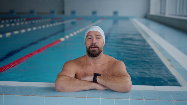 A mid adult man swimmer leaning on edge and looking at camera in public indoors swimming pool.