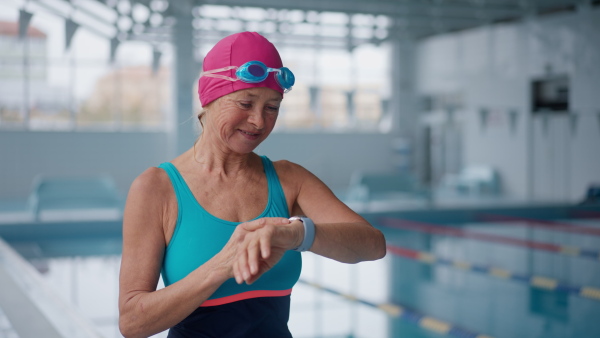 A senior woman looking at watch and putting on goggles before swim in indoors swimming pool.