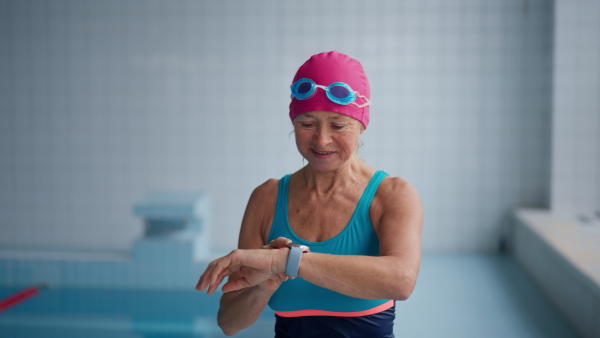 A senior woman looking at watch and putting on goggles before swim in indoors swimming pool.