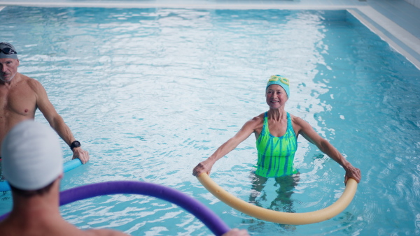 A therapist and senior woman and man doing aqua fitness with swim noodles indoors in swimming pool