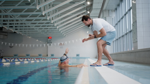 A senior swimmer man with his coach celebrating success indoors in swimming pool