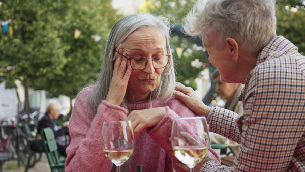 An unhappy senior woman sitting outdoors in cafe with her friend consoling her.