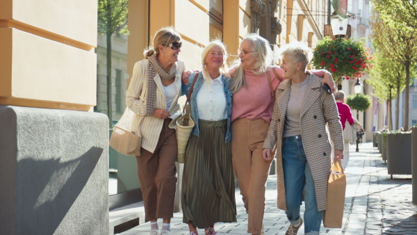 A group of cheerful senior women friends with shopping bags walking and holding together outdoors in city street.