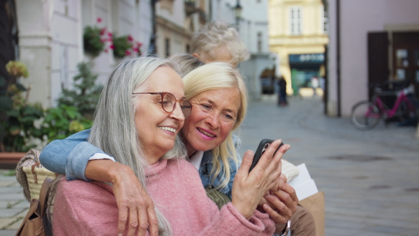 A group of cheerful senior women friends resting with smartphone in town.