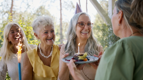 A senior woman getting small birthday cake from friends during celebration outdoors on patio.