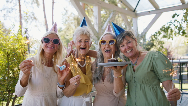 A senior woman getting small birthday cake from friends during celebration outdoors on patio, looking at camera.