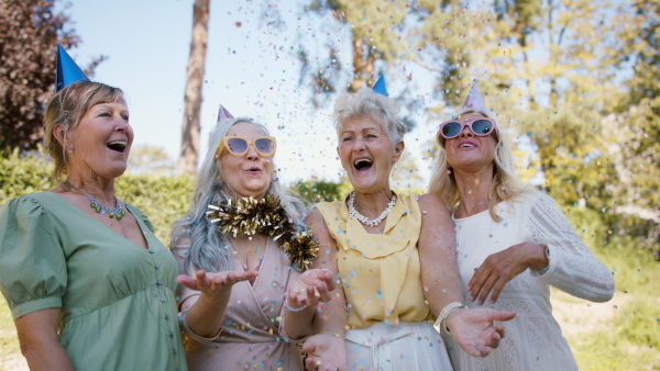 Happy senior women with party hats celebrating birthday, throwing confetti and hugging in the garden