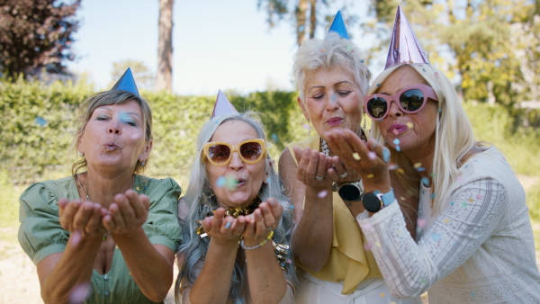 A close up of senior women with party hats celebrating birthday, blowing confetti and looking at camera in park