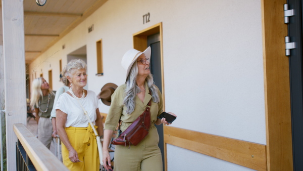 Senior women friends with suitcases greeting and embracing in front of hotel room outdoors on terrace.