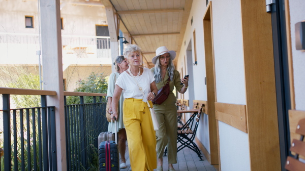 Senior women friends with suitcases greeting and embracing in front of hotel room outdoors on terrace.