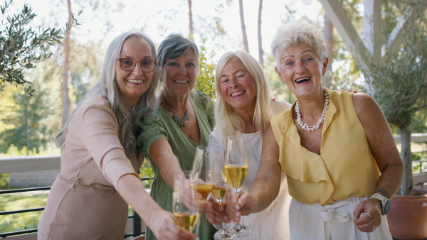 Happy senior women friends clinking glasses with champagne and looking at camera during celebration outdoors at terrace.