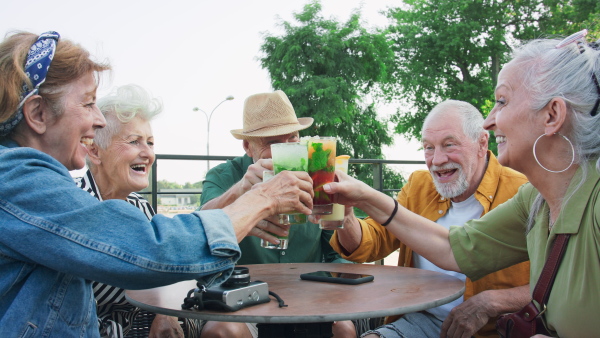 A group of happy senior friends tourists sitting outdoors in sidewalk cafe in city, enjoying drinks.