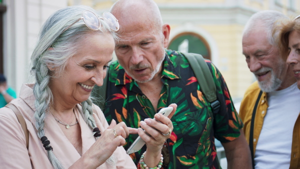 A group of happy senior friends tourists on a walk in city, using smartphone.