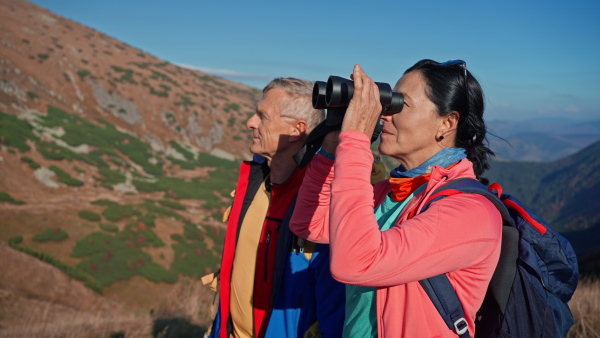 A senior couple hikers looking at view with binoculars together in mountains.