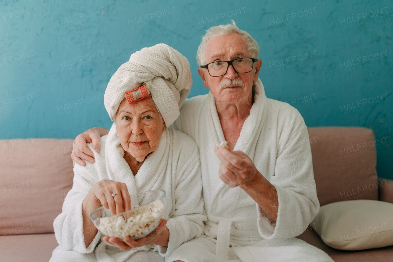 Happy senior couple sitting at a sofa in bathrobes and watching TV with popcorn.