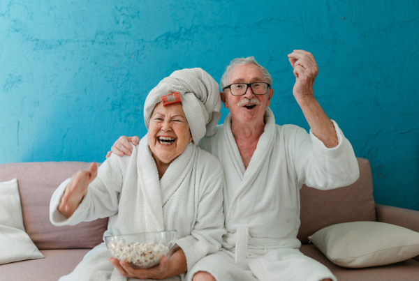 Happy senior couple sitting at a sofa in bathrobes and watching TV with popcorn.