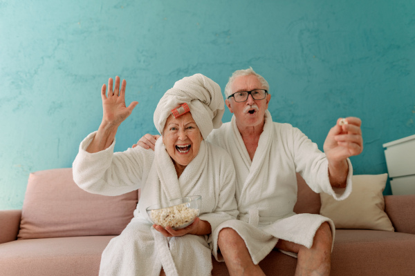 Happy senior couple sitting at a sofa in bathrobes and watching TV with popcorn.