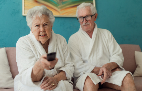 Happy senior couple sitting at a sofa in bathrobes and watching TV.