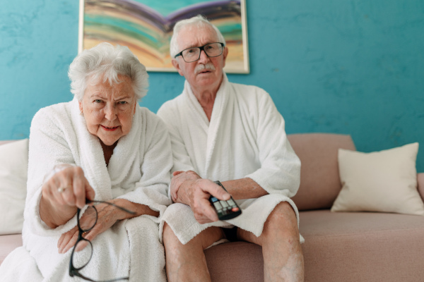 Happy senior couple sitting at a sofa in bathrobes and watching TV with popcorn.